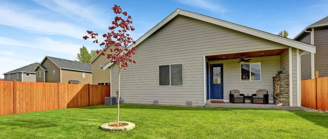 A residential home with a large fenced in back yard. The lawn has been neatly mowed and the small tree in the yard has been pruned.