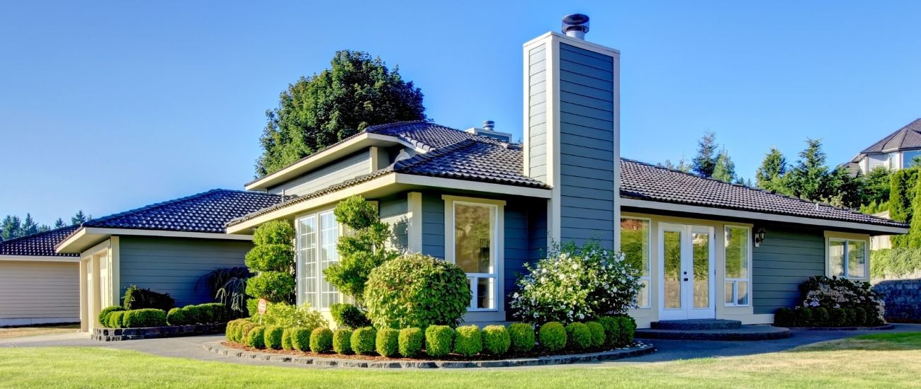 A residential home with blue siding. The surrounding lawn is neatly mowed and the landscape beds lining the home are filled with fresh mulch and neatly pruned shrubs and bushes.